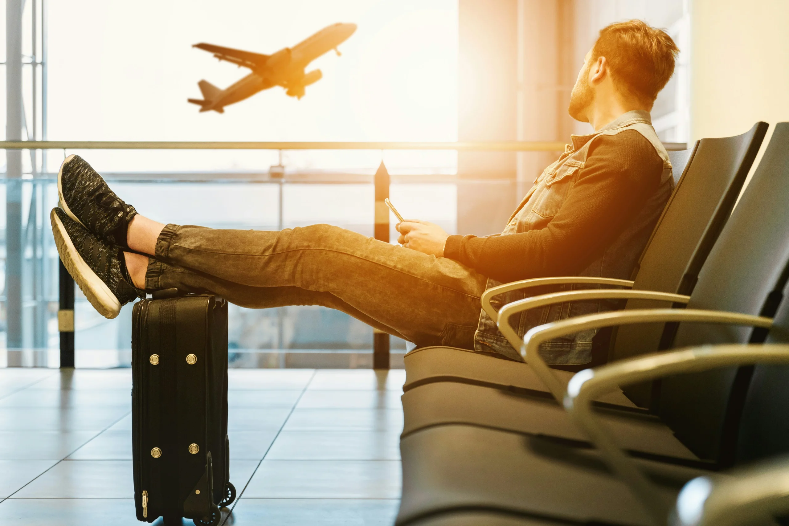 Relaxed man waiting at the airport with suitcase, watching a plane take off at sunset